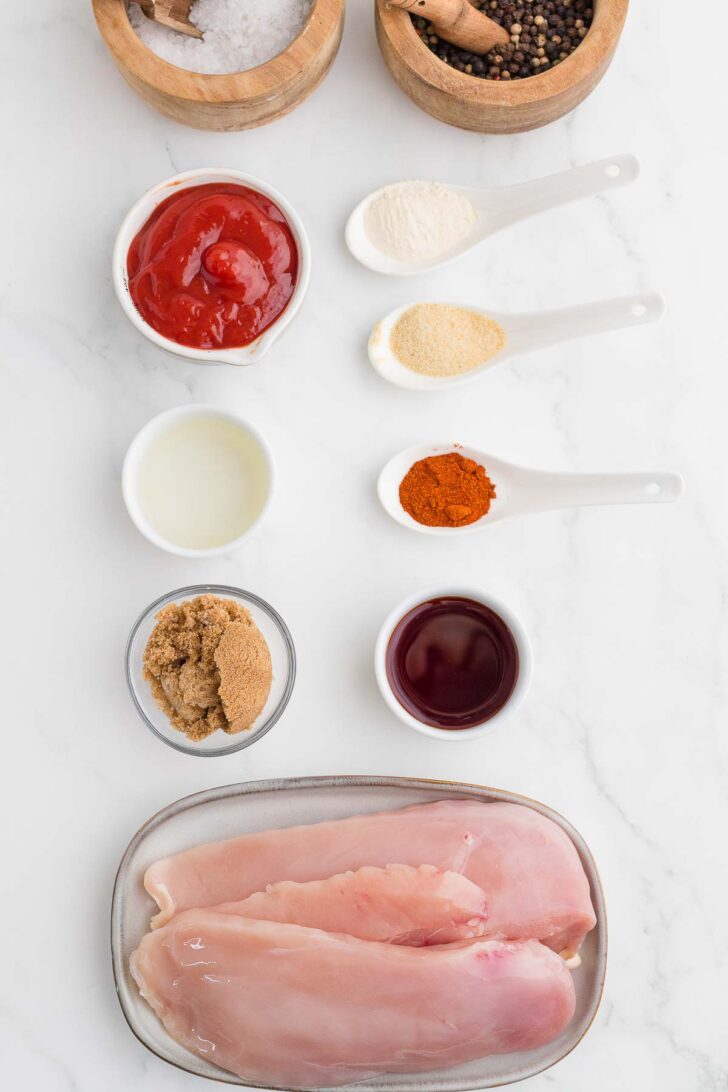 Flat lay of raw chicken breasts, ketchup, vinegar, brown sugar, onion powder, garlic powder, paprika, and spices in bowls and spoons on a white marble surface.
