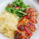 A plate with sliced barbecue chicken breast, mashed potatoes, and a fresh green salad with sliced tomatoes, garnished with chopped herbs. A fork rests on the side of the plate.