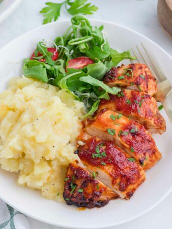 A plate with sliced barbecue chicken breast, mashed potatoes, and a fresh green salad with sliced tomatoes, garnished with chopped herbs. A fork rests on the side of the plate.