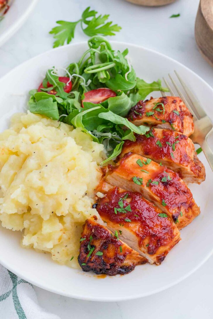 A plate with sliced barbecue chicken breast, mashed potatoes, and a fresh green salad with sliced tomatoes, garnished with chopped herbs. A fork rests on the side of the plate.