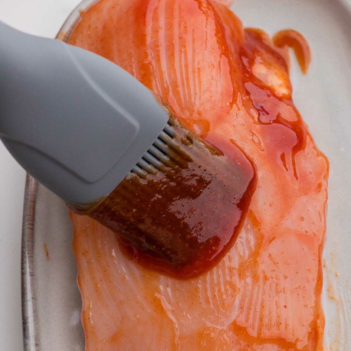 A silicone brush spreads barbecue sauce onto a raw chicken breast placed on a light-colored ceramic plate.