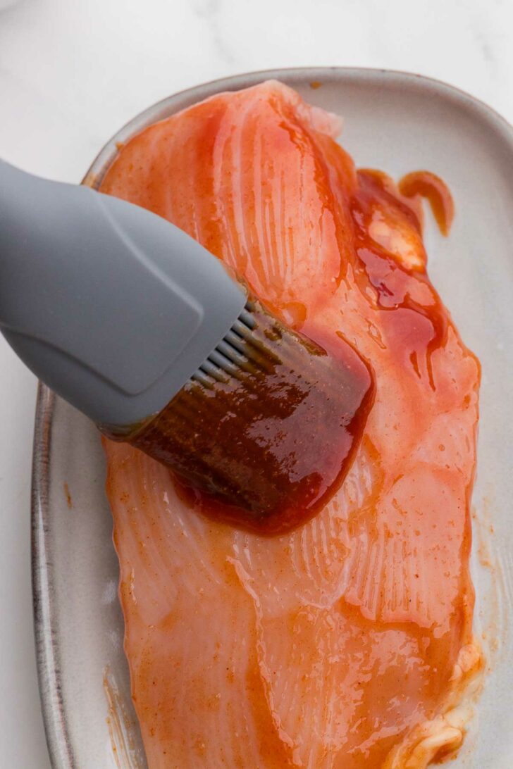 A silicone brush spreads barbecue sauce onto a raw chicken breast placed on a light-colored ceramic plate.