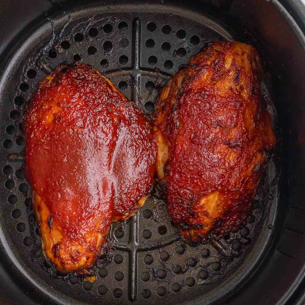 Two barbecued chicken breasts with a shiny glaze are cooking in a black air fryer basket.