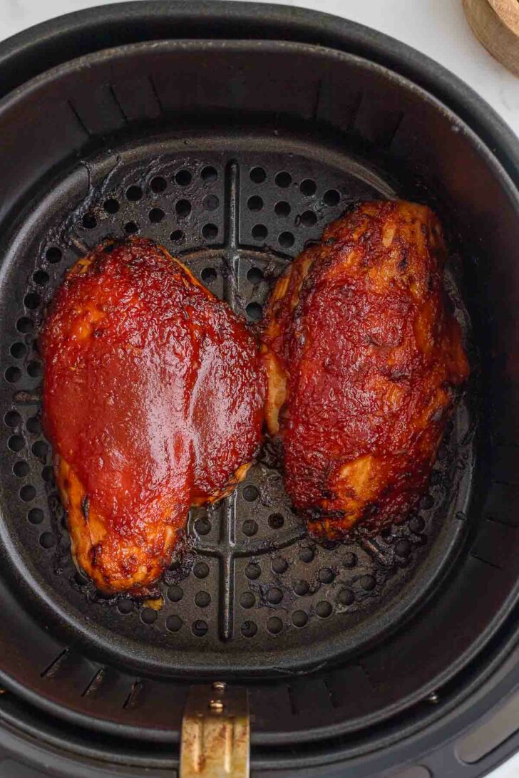 Two barbecued chicken breasts with a shiny glaze are cooking in a black air fryer basket.