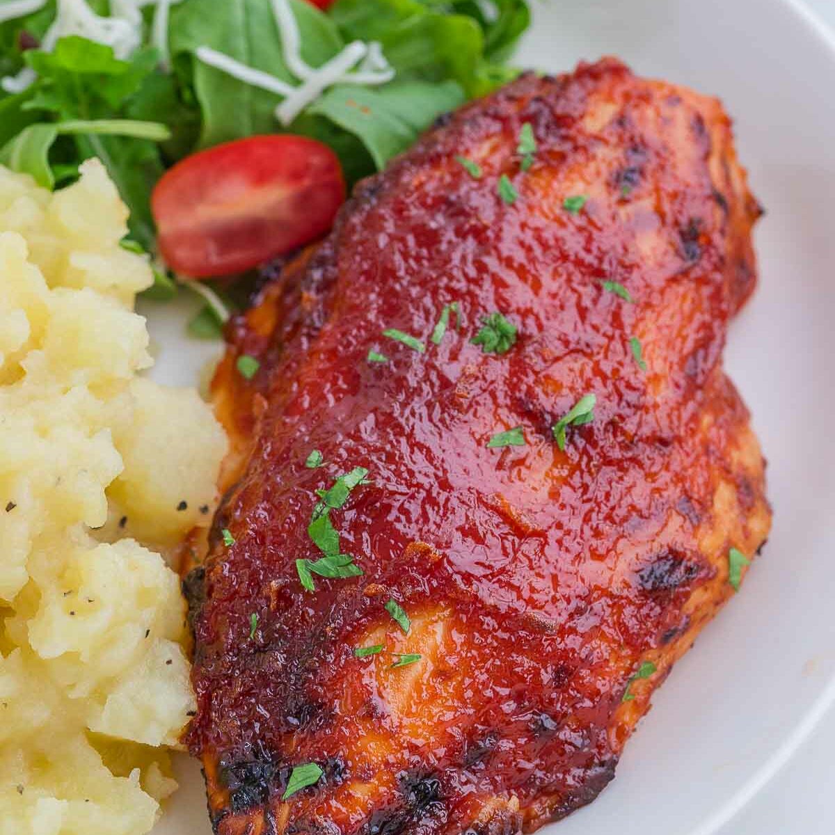 A plate with barbecue-glazed chicken breast, mashed potatoes, and a green salad with cherry tomatoes, garnished with chopped herbs.