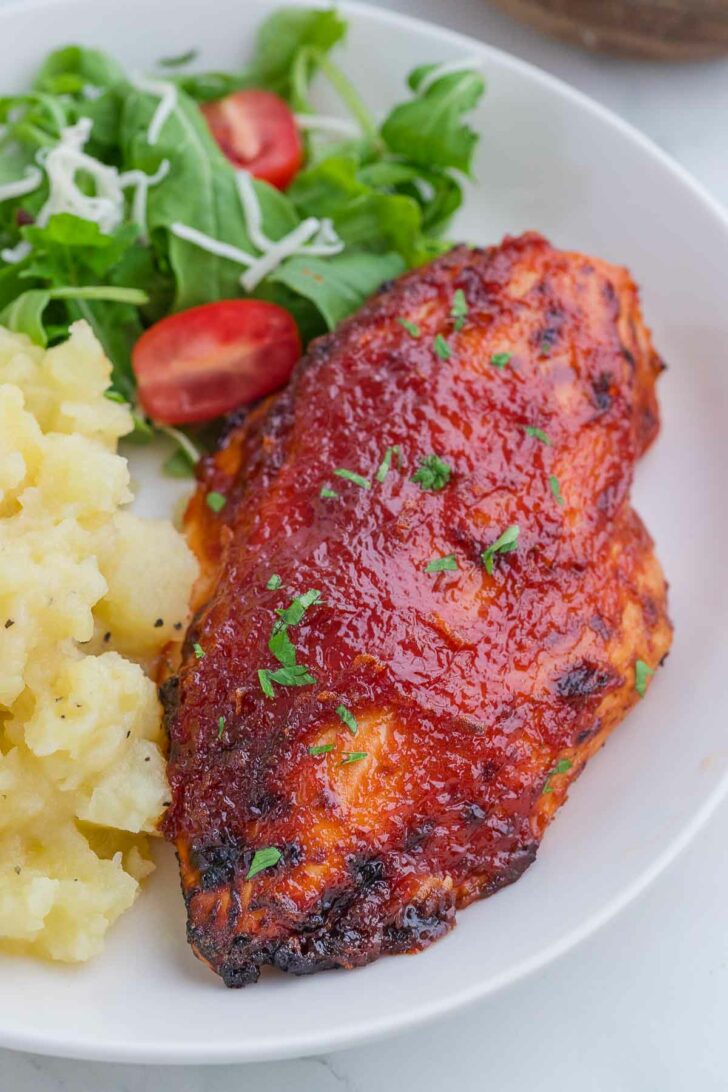 A plate with barbecue-glazed chicken breast, mashed potatoes, and a green salad with cherry tomatoes, garnished with chopped herbs.