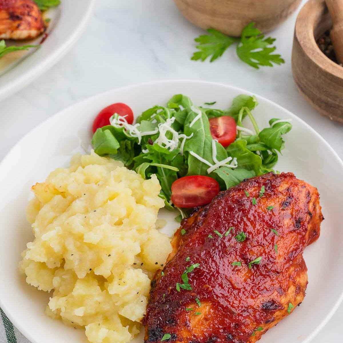 A plate with mashed potatoes, barbecue-glazed chicken breast, and a fresh salad with cherry tomatoes and shredded cheese. Wooden bowls with salt and pepper are in the background on a white surface.