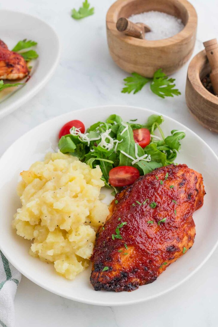 A plate with mashed potatoes, barbecue-glazed chicken breast, and a fresh salad with cherry tomatoes and shredded cheese. Wooden bowls with salt and pepper are in the background on a white surface.
