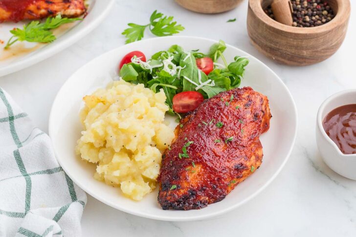 A white plate with barbecued chicken breast, mashed potatoes, and a green salad with cherry tomatoes and shredded cheese. A bowl of BBQ sauce, pepper grinder, and parsley garnish are nearby on a marble surface.