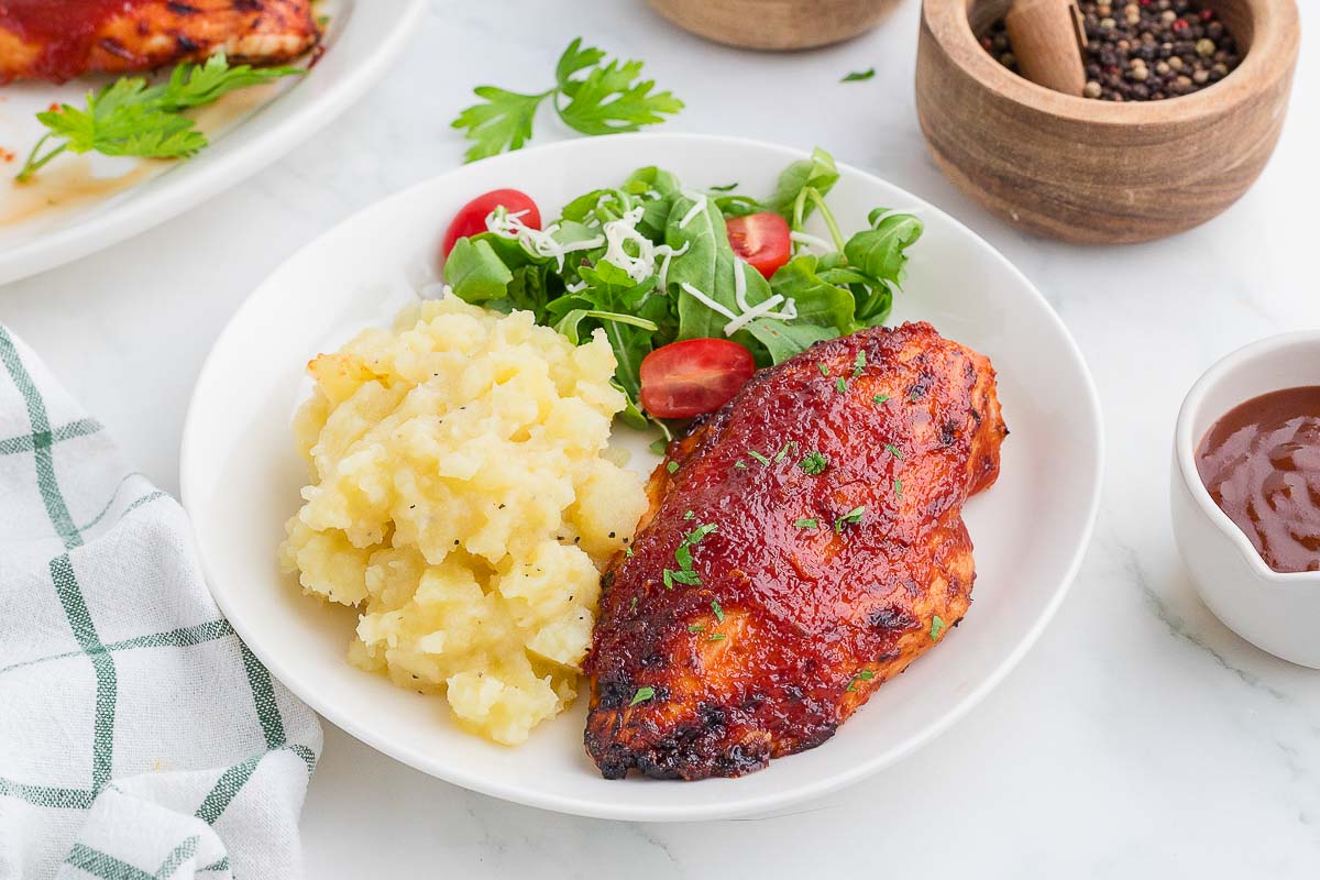 A white plate with barbecued chicken breast, mashed potatoes, and a green salad with cherry tomatoes and shredded cheese. A bowl of BBQ sauce, pepper grinder, and parsley garnish are nearby on a marble surface.