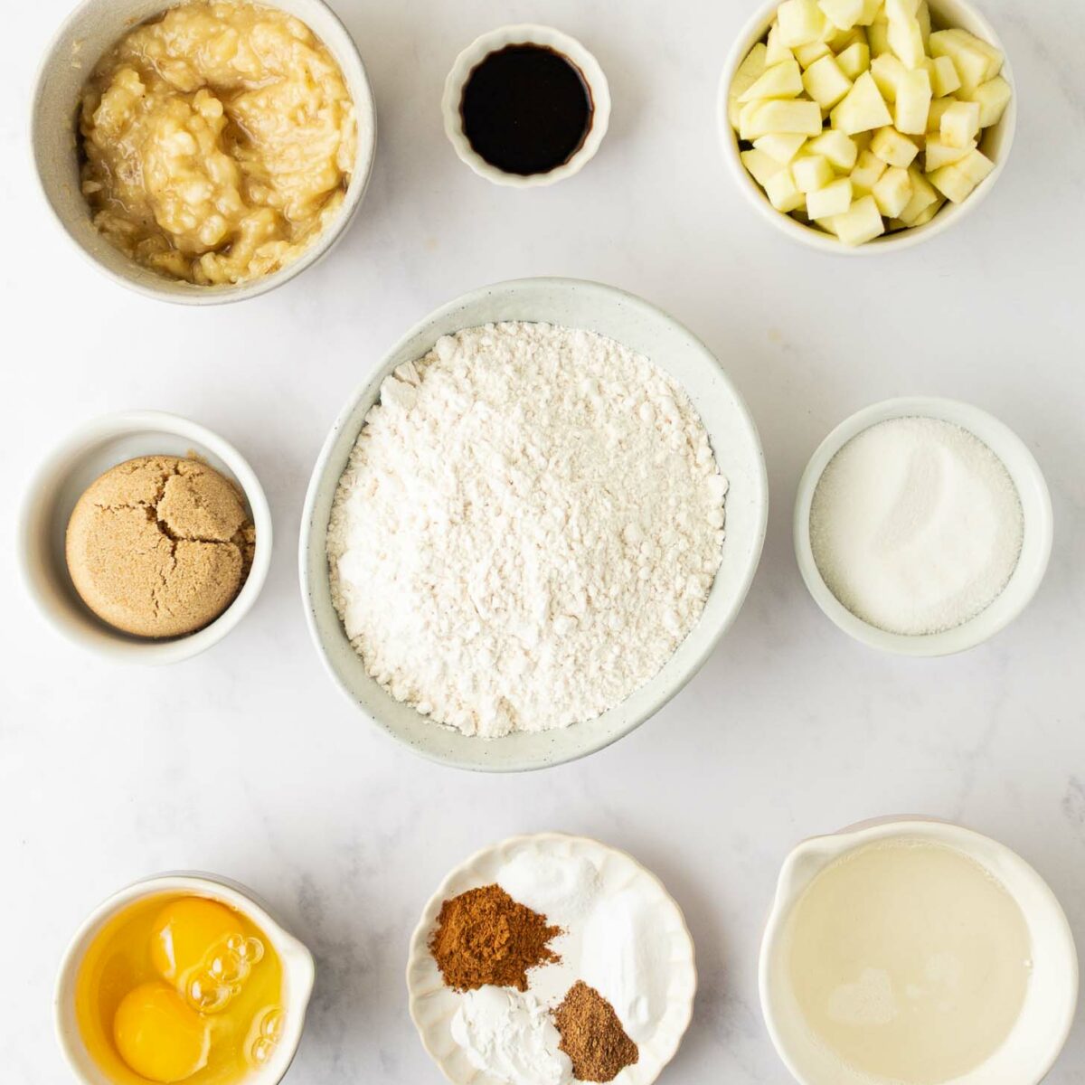 Bowls of baking ingredients arranged on a white surface, including mashed banana, vanilla, chopped apple, brown sugar, flour, white sugar, two eggs, spices, and milk.