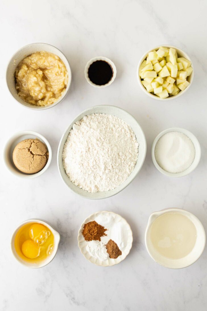 Bowls of baking ingredients arranged on a white surface, including mashed banana, vanilla, chopped apple, brown sugar, flour, white sugar, two eggs, spices, and milk.
