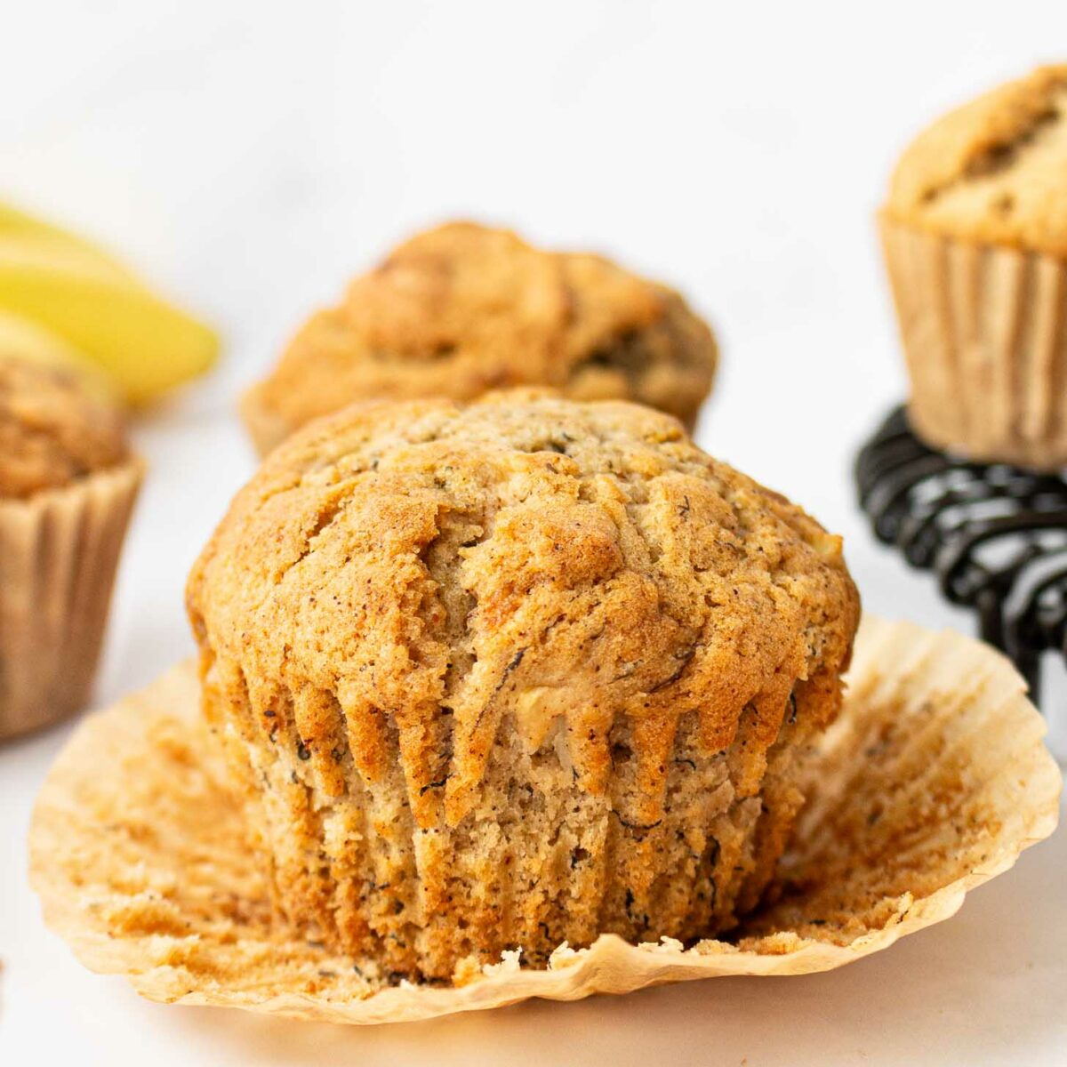 An apple banana muffin with its wrapper peeled back sits in the foreground, with other muffins and a sliced banana blurred in the background on a light surface.