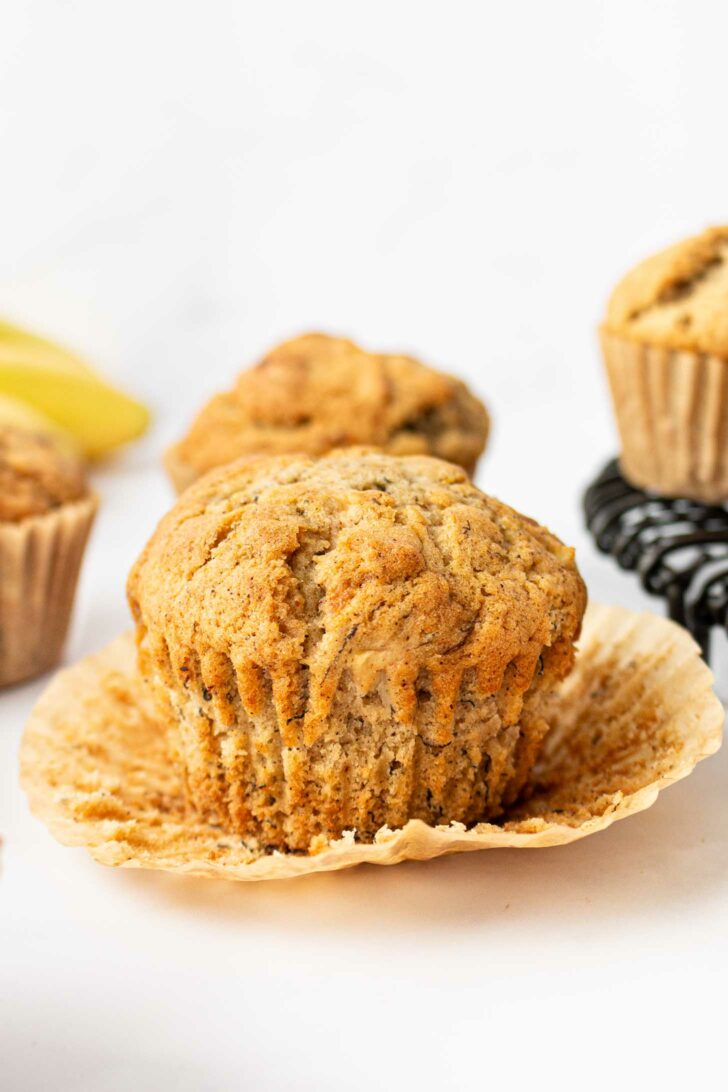 An apple banana muffin with its wrapper peeled back sits in the foreground, with other muffins and a sliced banana blurred in the background on a light surface.
