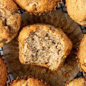 A close-up of several apple banana muffins on a cooling rack, with one muffin in the center unwrapped and partially eaten, showing its moist, crumbly interior.