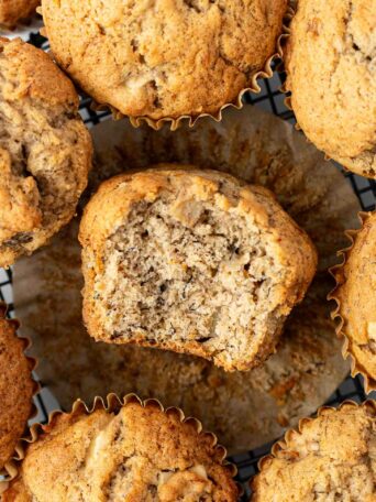 A close-up of several apple banana muffins on a cooling rack, with one muffin in the center unwrapped and partially eaten, showing its moist, crumbly interior.