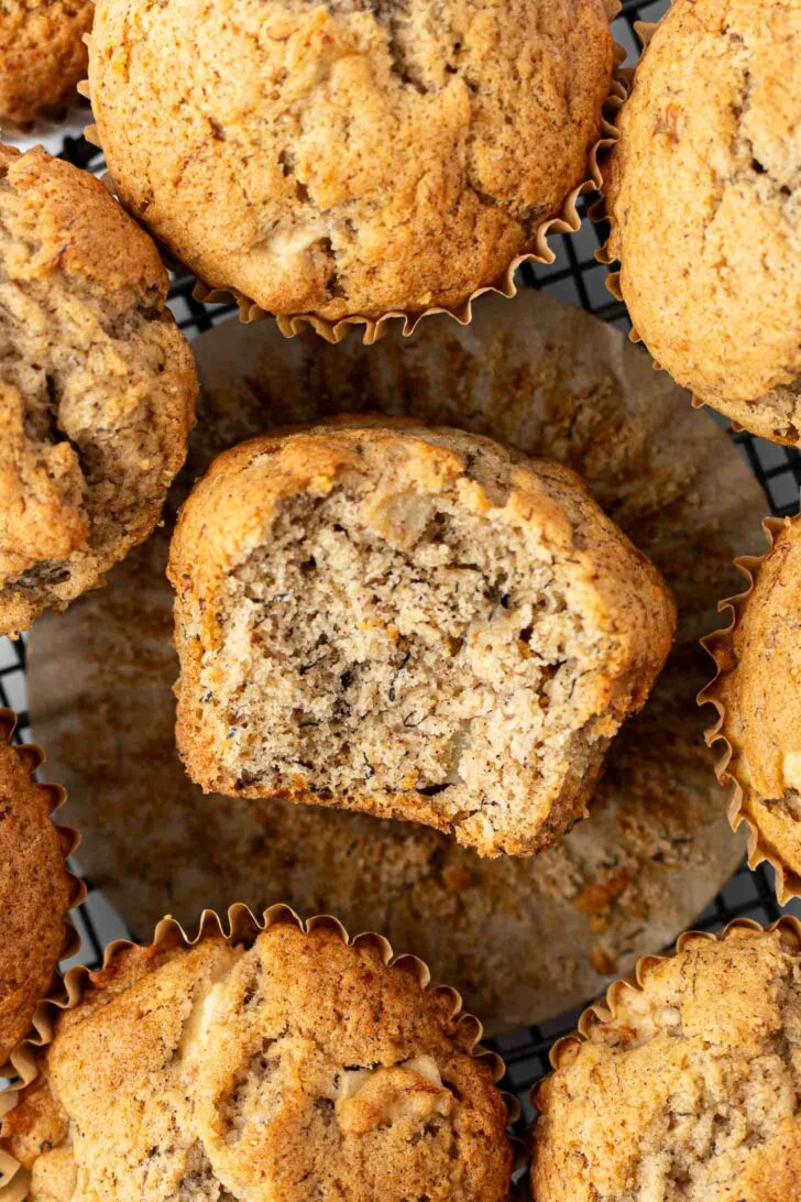 A close-up of several apple banana muffins on a cooling rack, with one muffin in the center unwrapped and partially eaten, showing its moist, crumbly interior.