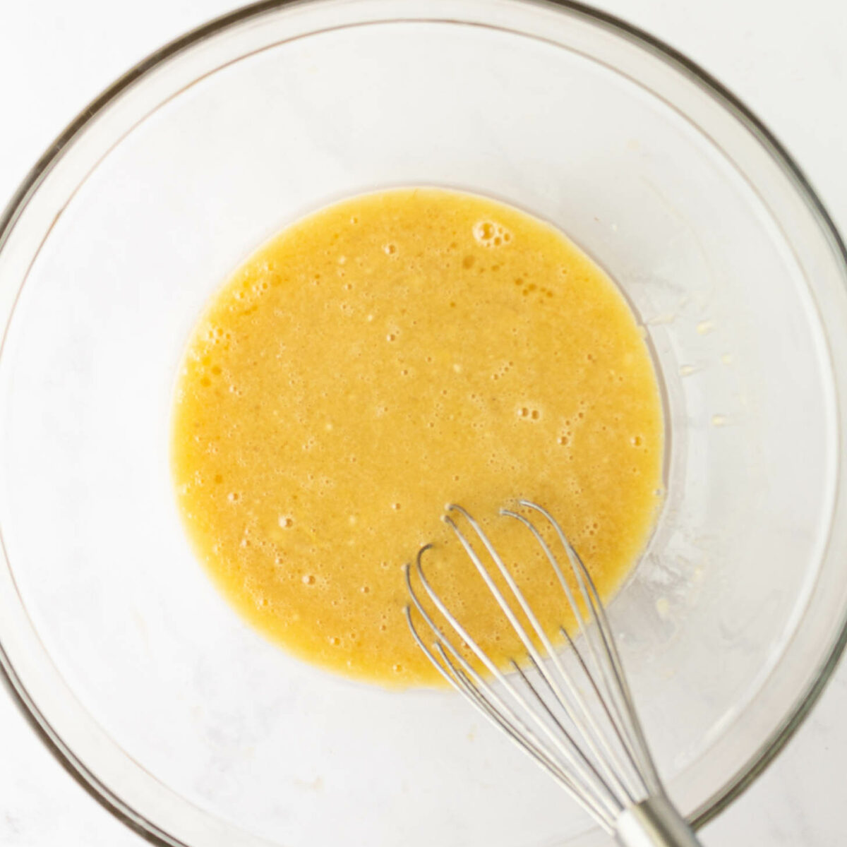 A glass bowl containing a yellow, whisked liquid mixture, with a metal whisk partially visible in the lower right corner. The background is a white countertop.