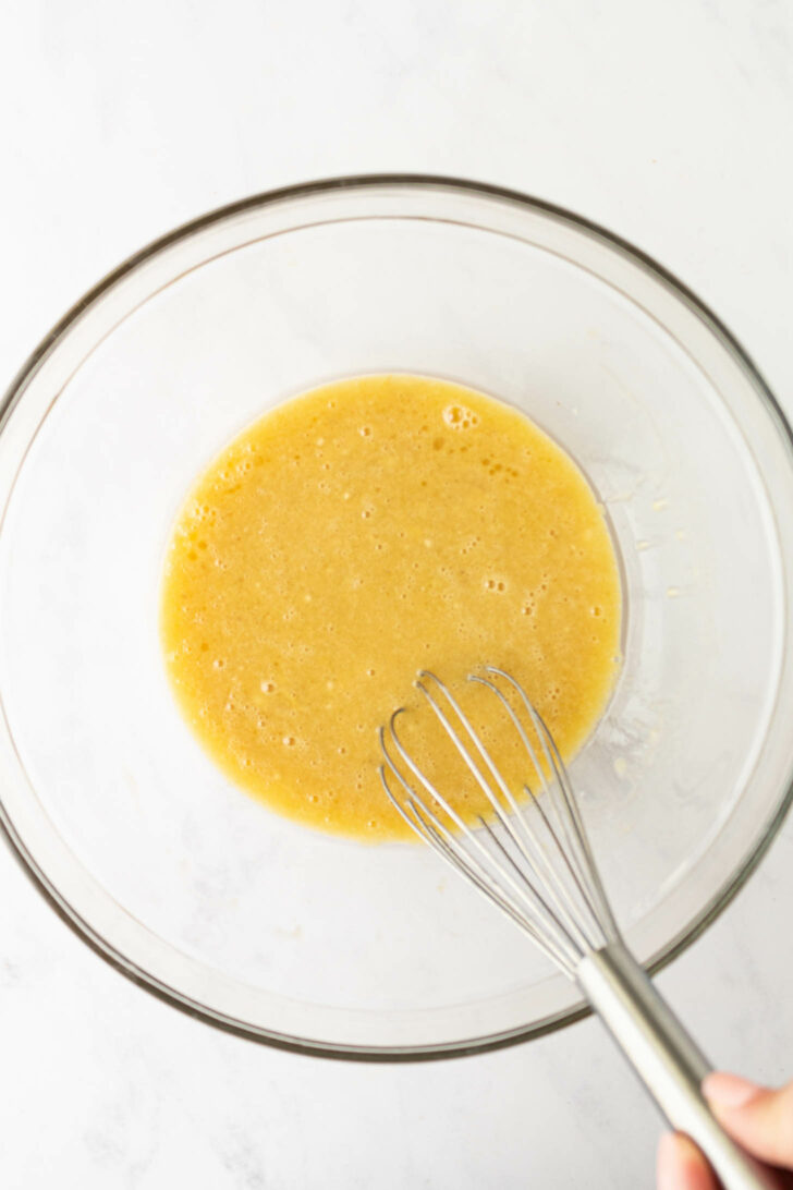 A glass bowl containing a yellow, whisked liquid mixture, with a metal whisk partially visible in the lower right corner. The background is a white countertop.