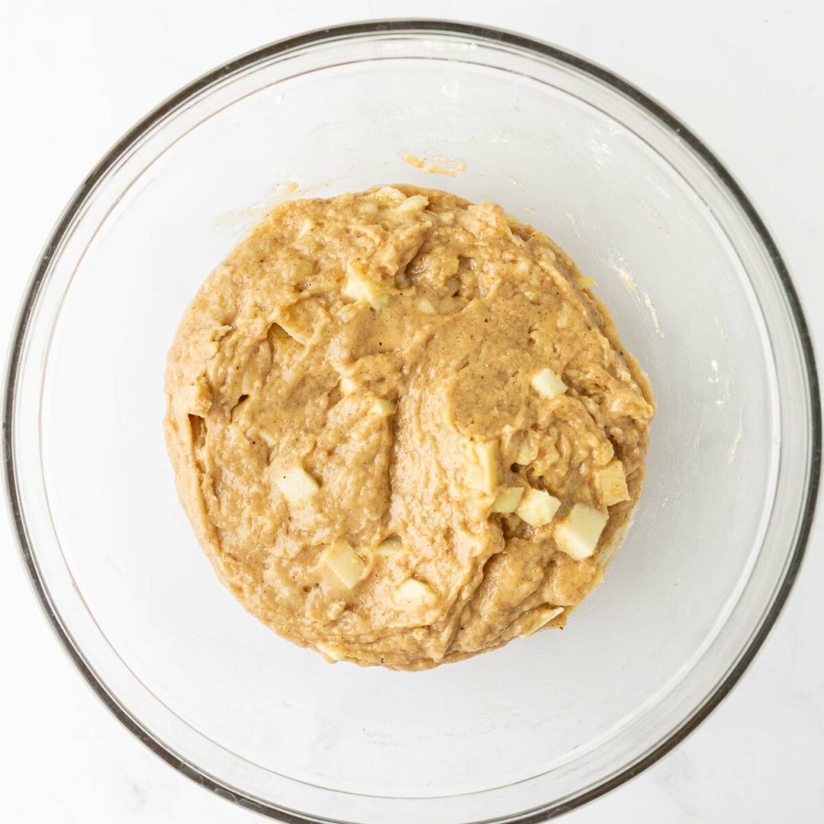 A glass bowl filled with thick, light brown batter mixed with small chunks of apple and banana, sitting on a white surface.