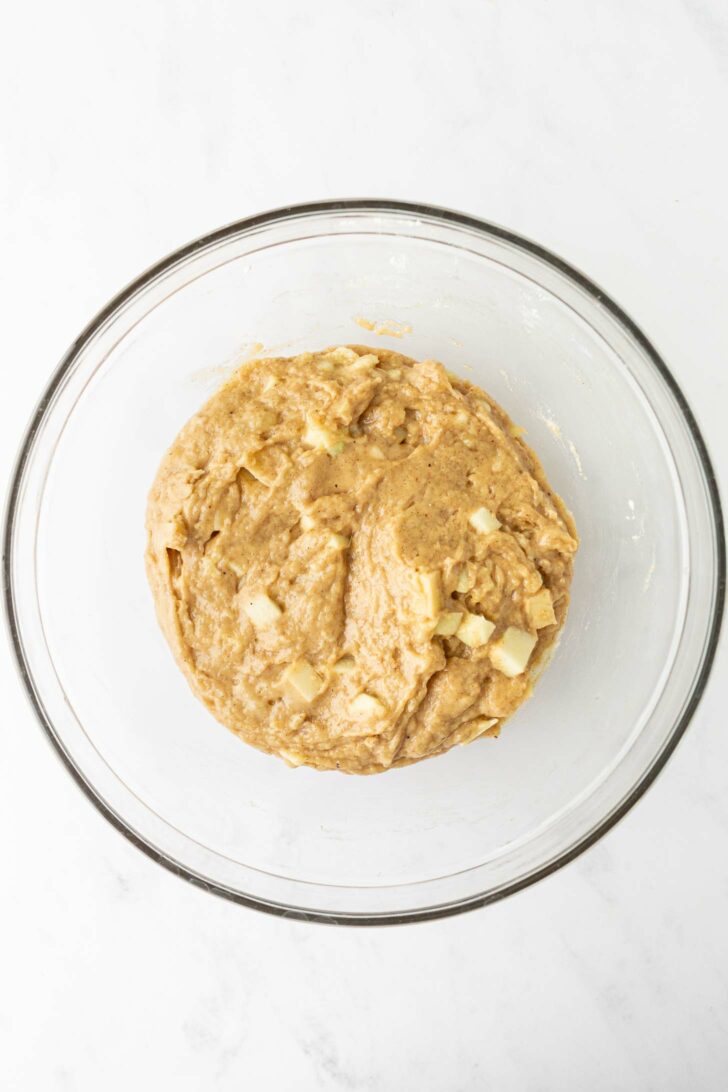 A glass bowl filled with thick, light brown batter mixed with small chunks of apple and banana, sitting on a white surface.