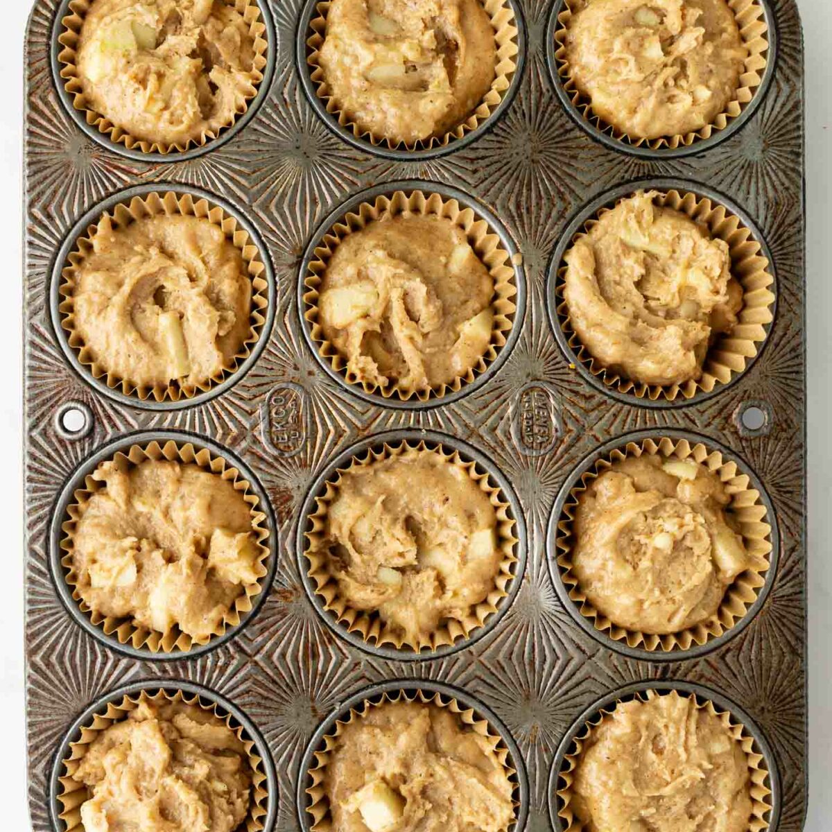 A muffin tin with twelve wells, each lined with paper and filled with unbaked muffin batter containing visible chunks, resting on a white surface.