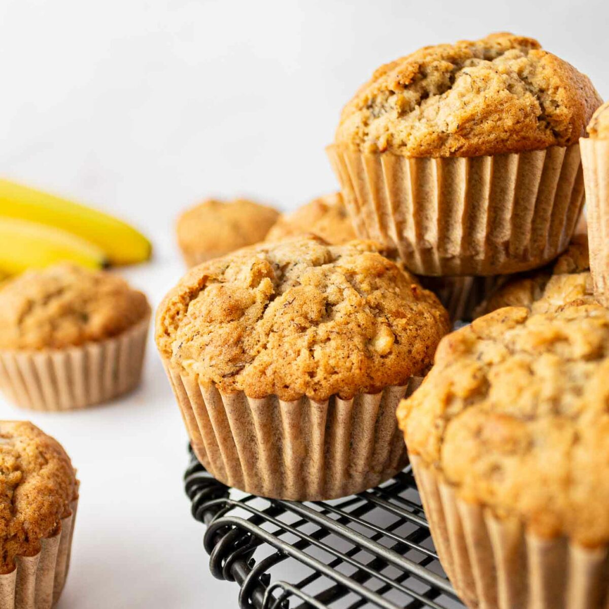Freshly baked apple banana muffins in paper liners are stacked on a black wire cooling rack, with ripe bananas in the background. The muffins are golden brown with a textured, crumbly top.