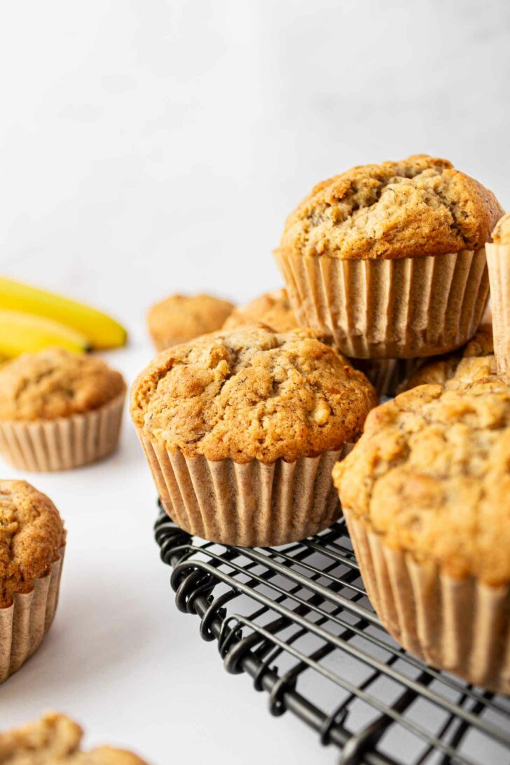 Freshly baked apple banana muffins in paper liners are stacked on a black wire cooling rack, with ripe bananas in the background. The muffins are golden brown with a textured, crumbly top.