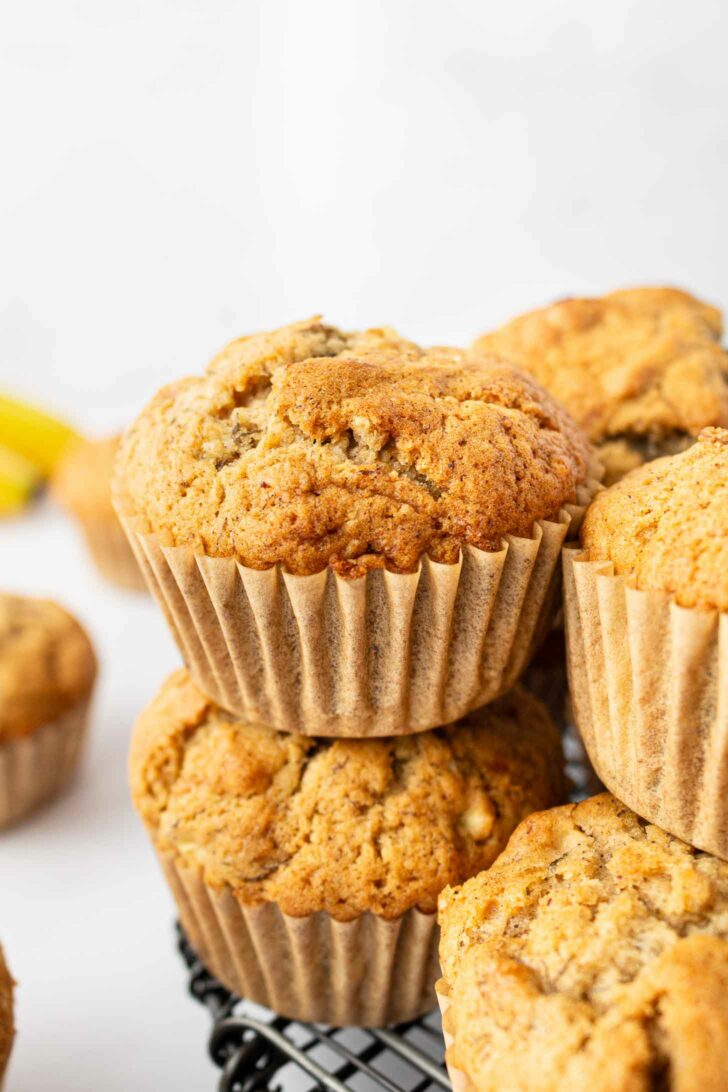 Close-up of golden brown muffins stacked on a black wire rack, with a light background and a few more muffins visible in the background.