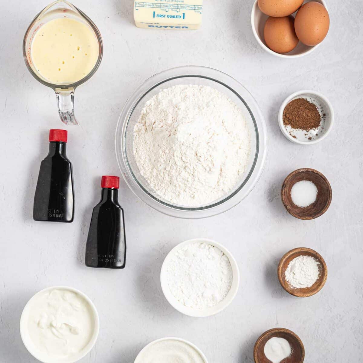 A flat lay of baking ingredients including flour, sugar, eggs, butter sticks, vanilla extract, sour cream, baking soda, salt, baking powder, eggnog, and a small bowl of spices, arranged on a light gray surface.