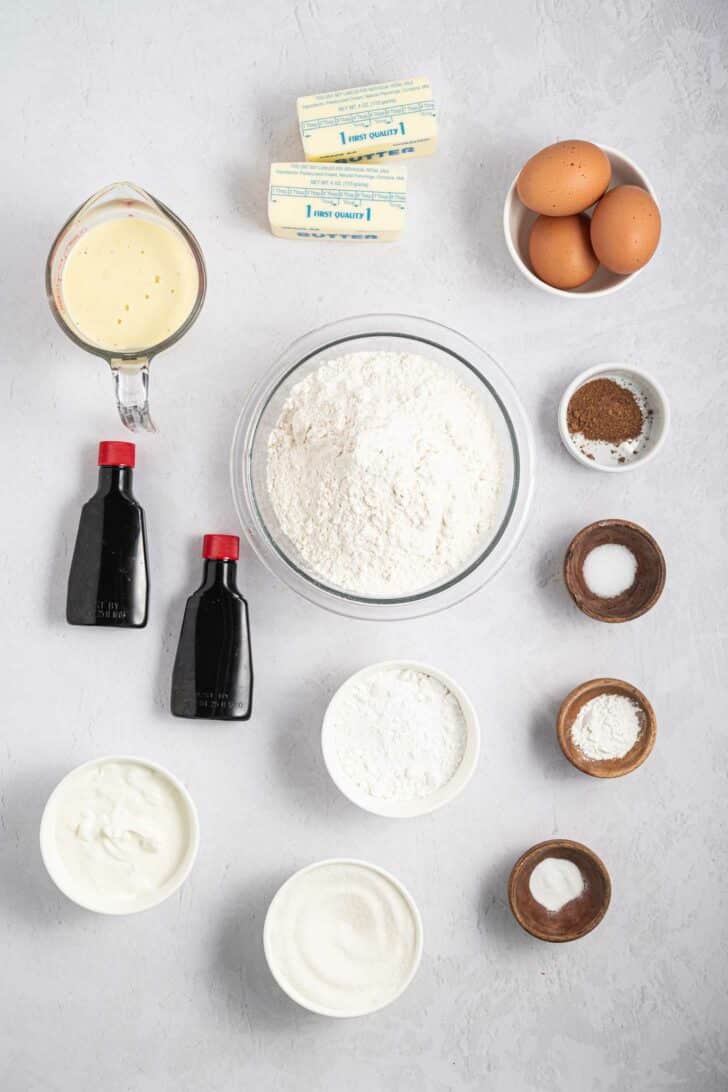A flat lay of baking ingredients including flour, sugar, eggs, butter sticks, vanilla extract, sour cream, baking soda, salt, baking powder, eggnog, and a small bowl of spices, arranged on a light gray surface.
