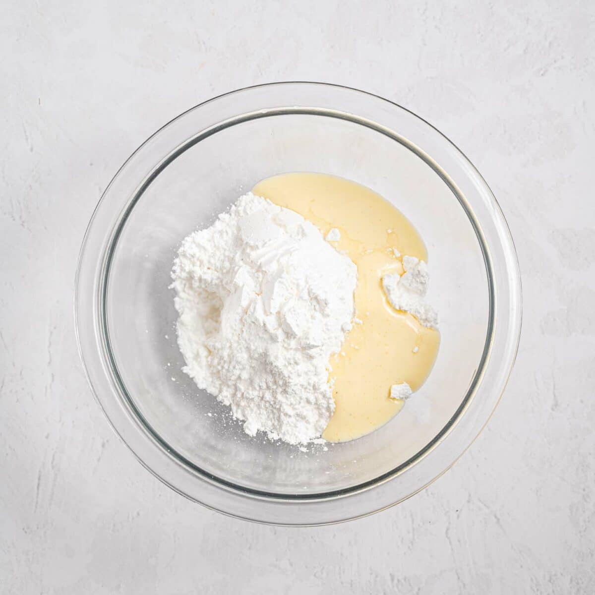 A glass mixing bowl containing a mound of white powdered sugar and a pool of pale yellow liquid, set on a light textured surface.