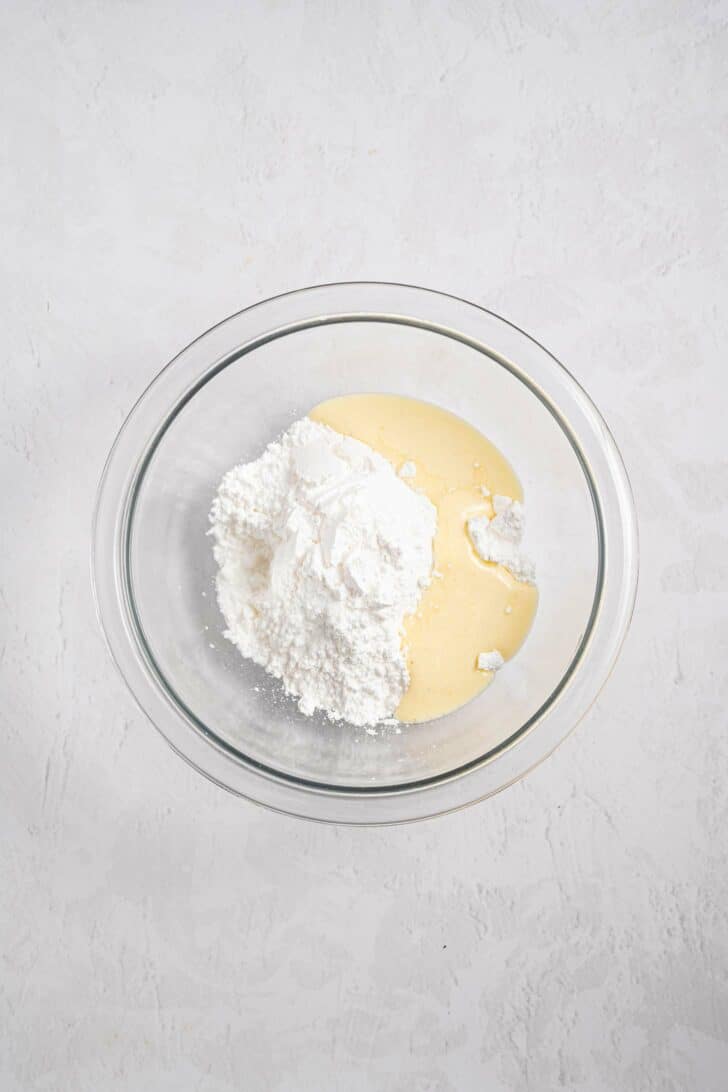 A glass mixing bowl containing a mound of white powdered sugar and a pool of pale yellow liquid, set on a light textured surface.