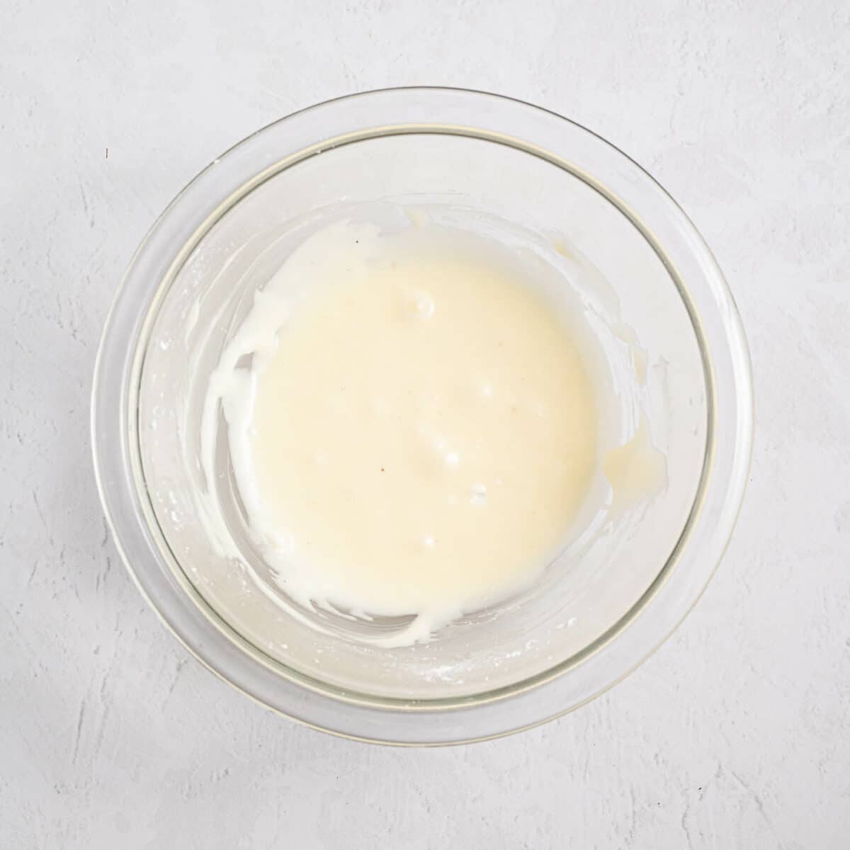A clear glass bowl containing a small amount of smooth, white batter or sauce sits on a light, textured surface.