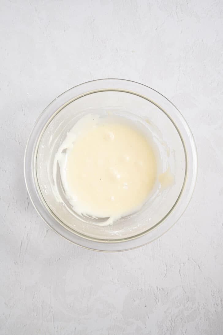 A clear glass bowl containing a small amount of smooth, white batter or sauce sits on a light, textured surface.