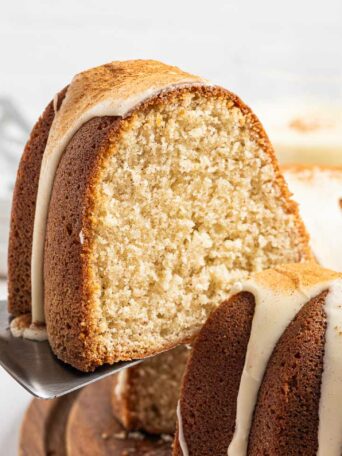 A close-up of a sliced bundt cake with white icing drizzled on top. A cake server lifts one slice, showing the cake’s moist, fluffy interior. The cake sits on a wooden board, with plates and utensils in the background.