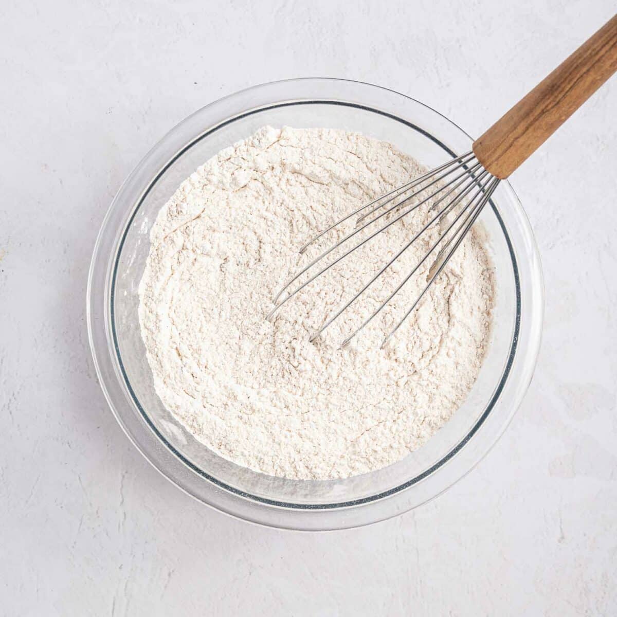 A glass bowl filled with flour mixture is being stirred with a metal whisk that has a wooden handle, set on a light-colored surface.