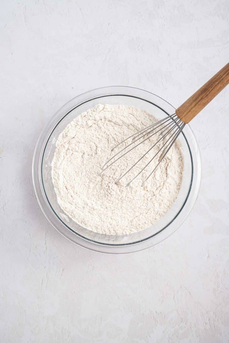 A glass bowl filled with flour mixture is being stirred with a metal whisk that has a wooden handle, set on a light-colored surface.