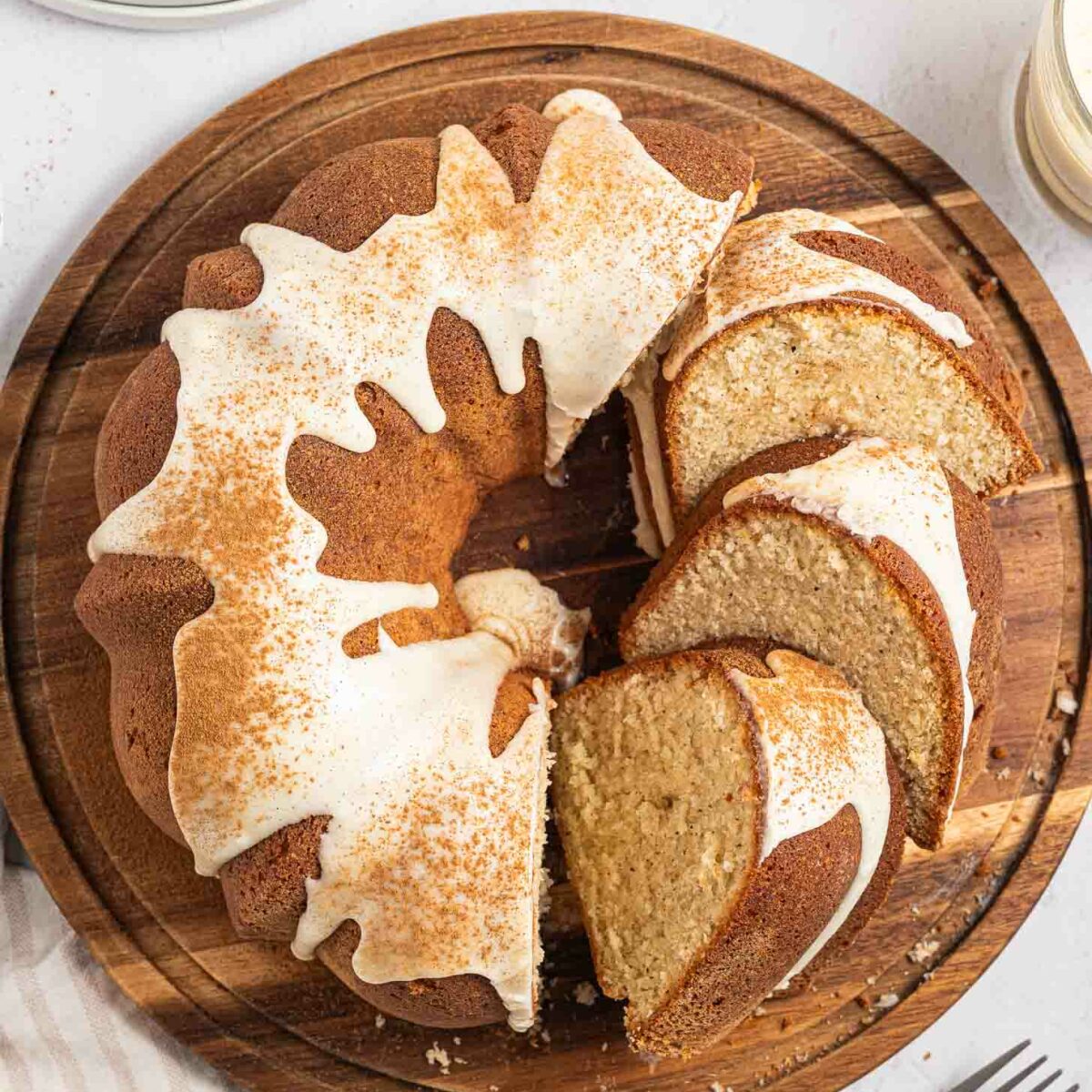 A sliced eggnog pound cake with white icing and a sprinkle of cinnamon sits on a round wooden board, with a few slices cut and separated. Forks and glasses of creamy drinks are nearby on a light striped cloth.