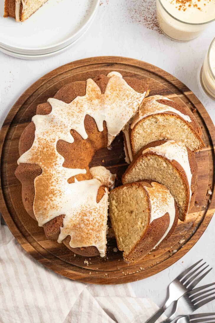 A sliced eggnog pound cake with white icing and a sprinkle of cinnamon sits on a round wooden board, with a few slices cut and separated. Forks and glasses of creamy drinks are nearby on a light striped cloth.