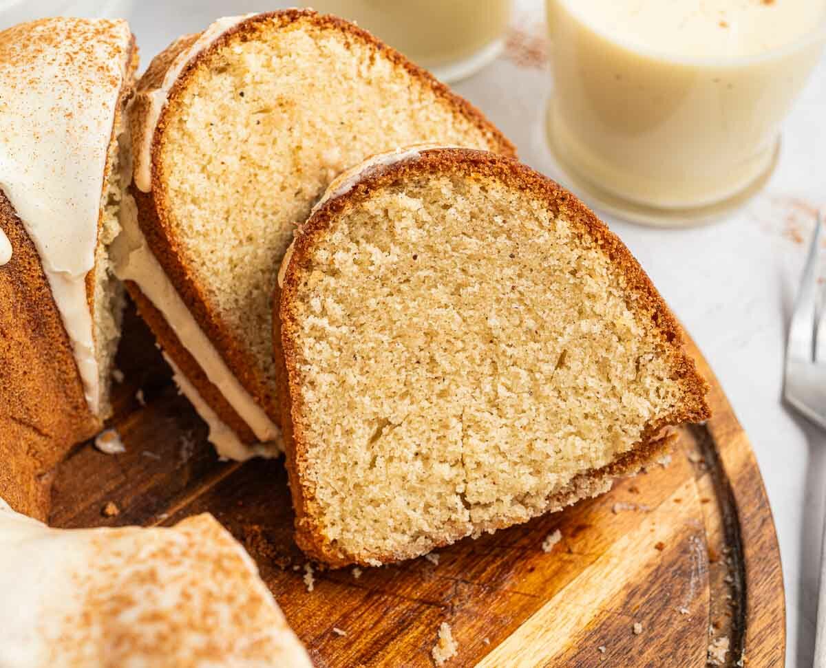 A close-up of sliced bundt cake with white icing, arranged on a wooden board. Two forks and glasses of creamy eggnog are in the background. The cake is moist with a light golden brown crumb.