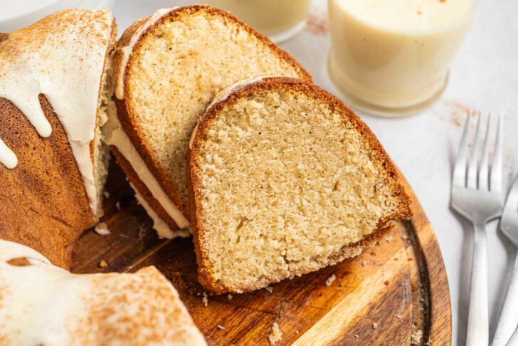 A close-up of sliced bundt cake with white icing, arranged on a wooden board. Two forks and glasses of creamy eggnog are in the background. The cake is moist with a light golden brown crumb.