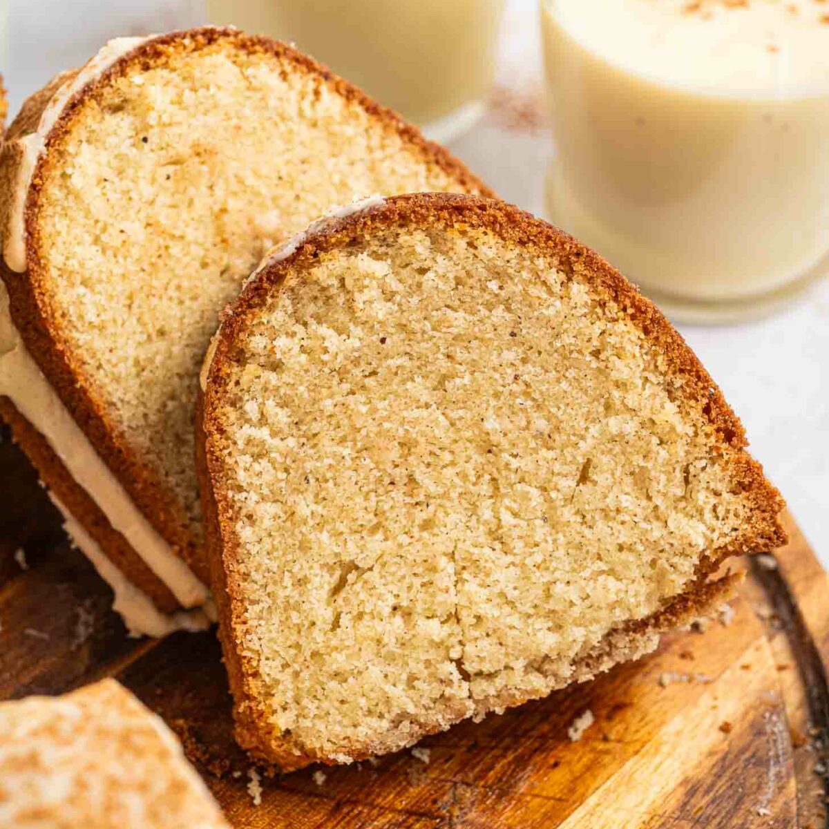 A close-up of a slice of bundt cake on a wooden board, with two glasses of eggnog topped with a sprinkle of spice in the background.