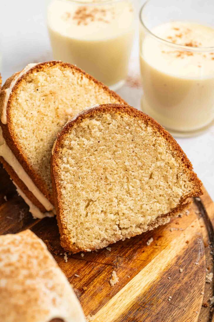 A close-up of a slice of bundt cake on a wooden board, with two glasses of eggnog topped with a sprinkle of spice in the background.