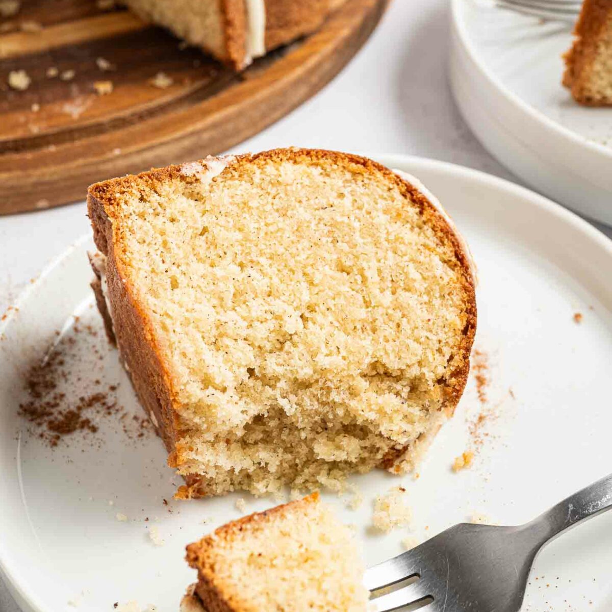 A close-up of a slice of moist eggnog pound cake on a white plate, with a fork holding a bite-sized piece. More cake slices and crumbs are in the background on another plate and a wooden board.