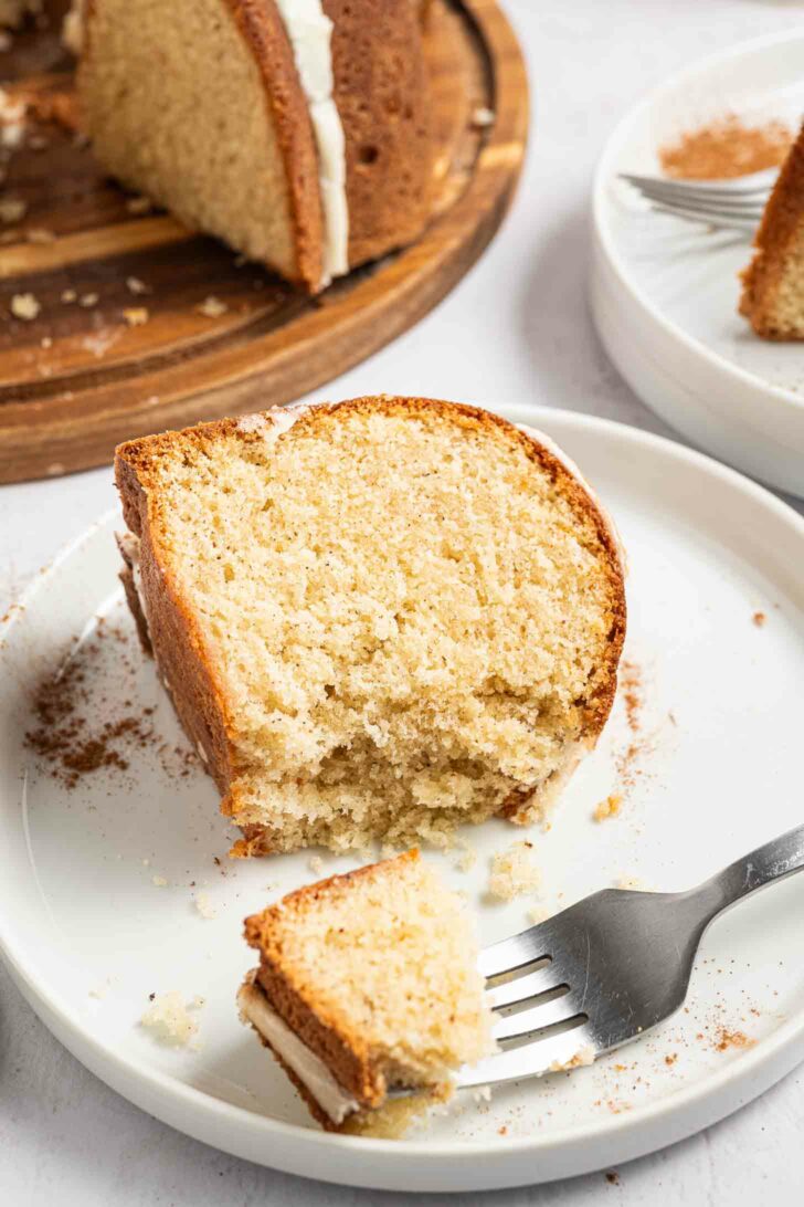 A close-up of a slice of moist eggnog pound cake on a white plate, with a fork holding a bite-sized piece. More cake slices and crumbs are in the background on another plate and a wooden board.