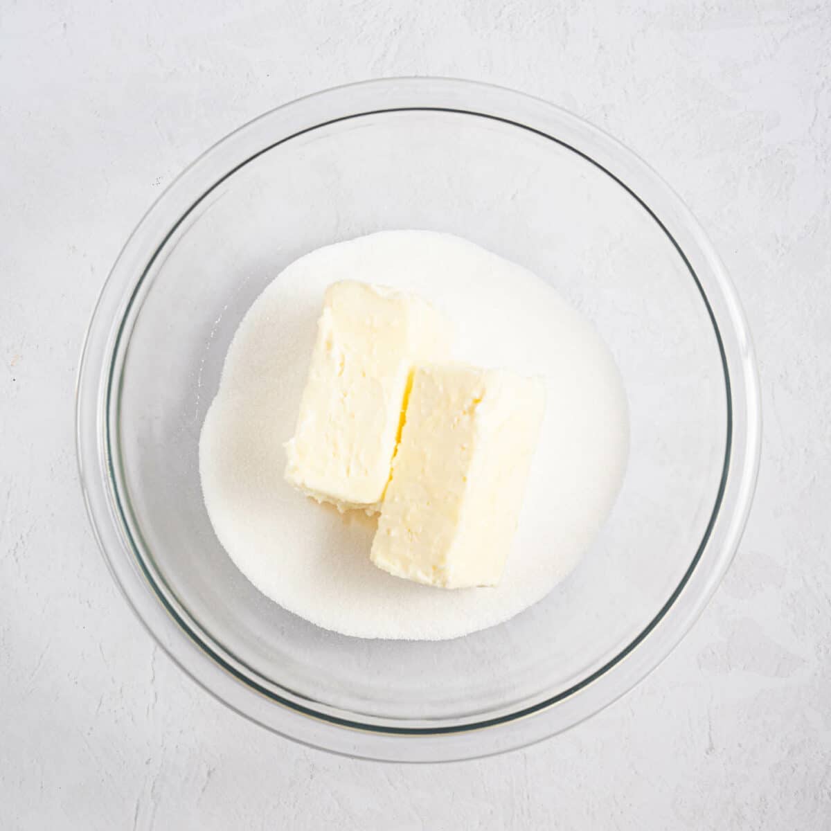 A glass bowl containing two sticks of butter placed on top of a pile of granulated white sugar, set on a light textured surface.