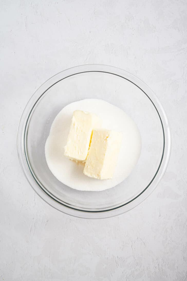 A glass bowl containing two sticks of butter placed on top of a pile of granulated white sugar, set on a light textured surface.
