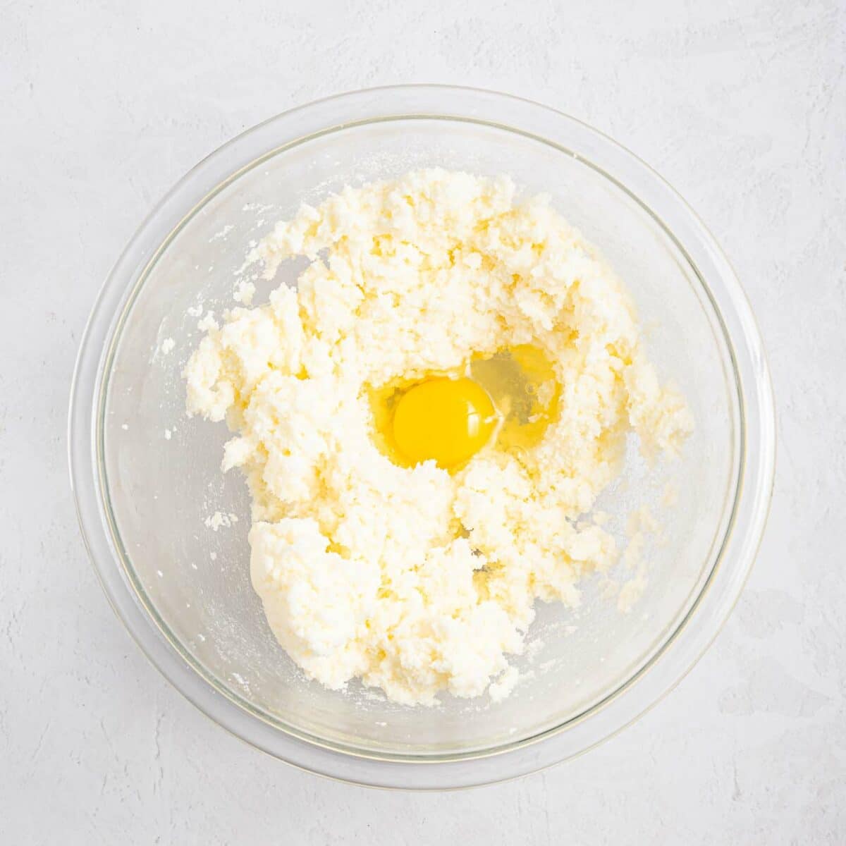 A glass mixing bowl with creamed butter and sugar, and a raw egg cracked on top, sitting on a light textured surface.