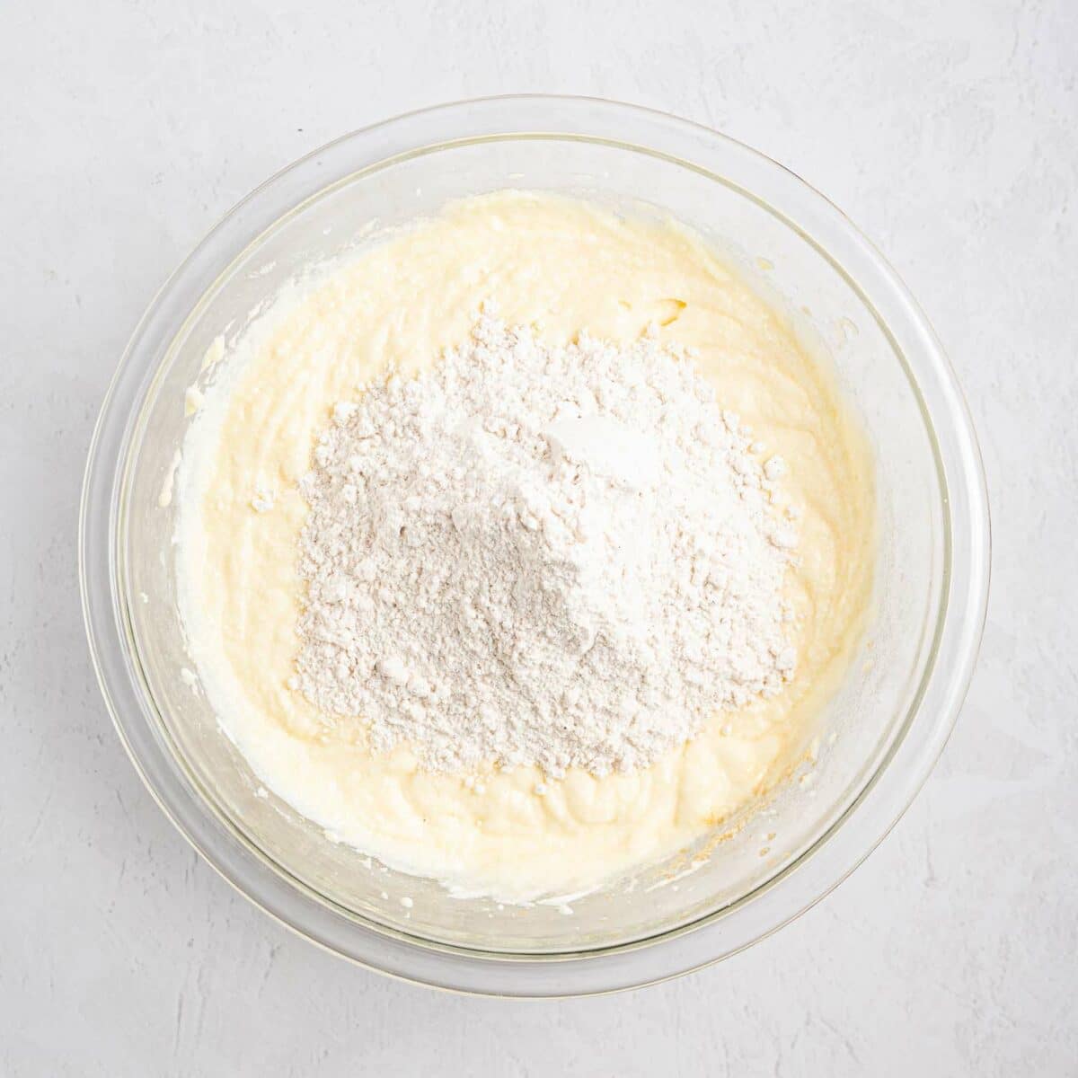 A glass bowl containing a creamy yellow batter with a mound of white flour on top, ready to be mixed, sits on a light textured surface.